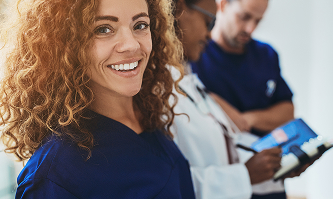 A smiling female healthcare worker with curly hair in blue scrubs, with two medical colleagues working in the background.