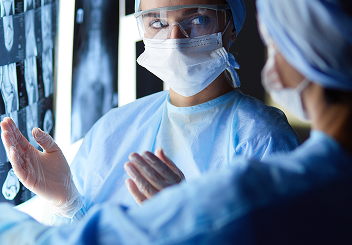 Two surgeons in surgical gowns, masks, and protective eyewear discussing X-ray scans mounted on a lightbox.