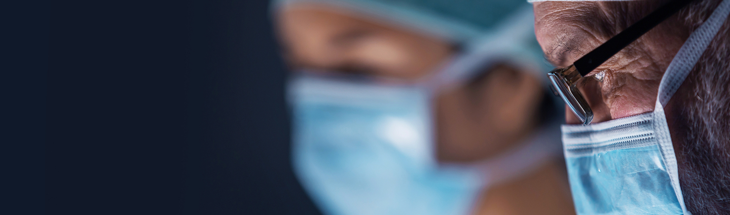 Close-up of a surgeon wearing glasses, a surgical mask, and a cap, looking down intently during a procedure.