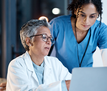 A doctor with short grey hair and glasses working at a computer alongside a colleague in blue scrubs.