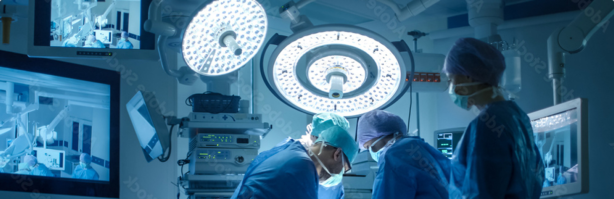 Surgical team operating under bright overhead surgical lights surrounded by medical monitors in a modern operating room