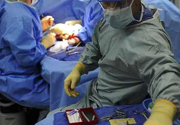 A surgeon in protective gown, mask, and safety goggles reaching toward surgical instruments on a tray, with doctors operating in the background.
