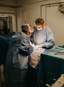 Two surgeons wearing blue gowns, face masks, and caps bent over a surgical table performing a procedure next to a tray of medical instruments.