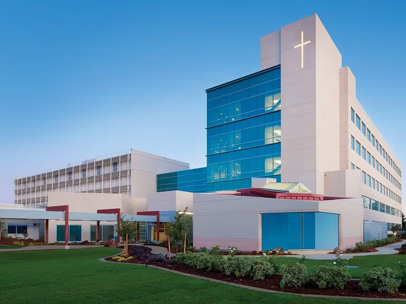 Exterior of a modern medical center building with a prominent cross on its upper tower and blue glass windows under a clear sky.