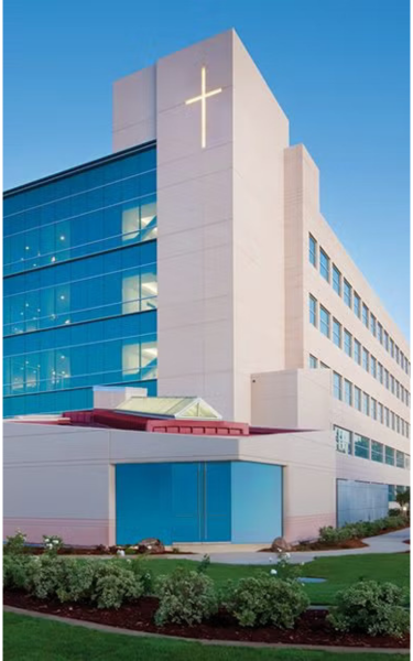 Exterior of a modern medical center building with a prominent cross on its upper tower and blue glass windows under a clear sky.