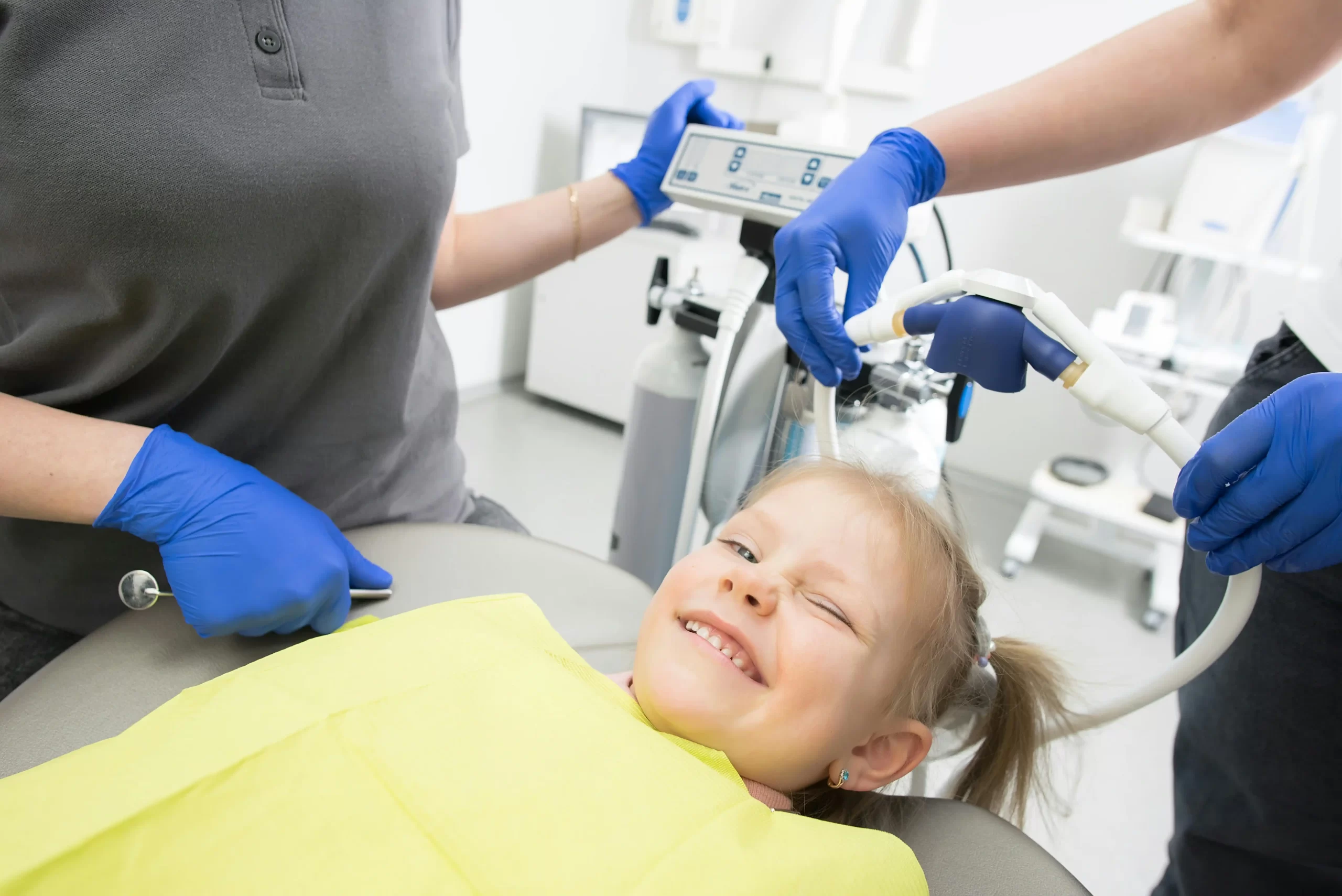 A smiling young girl in a dental chair as practitioners fit a nitrous oxide mask over her nose before a dental procedure.