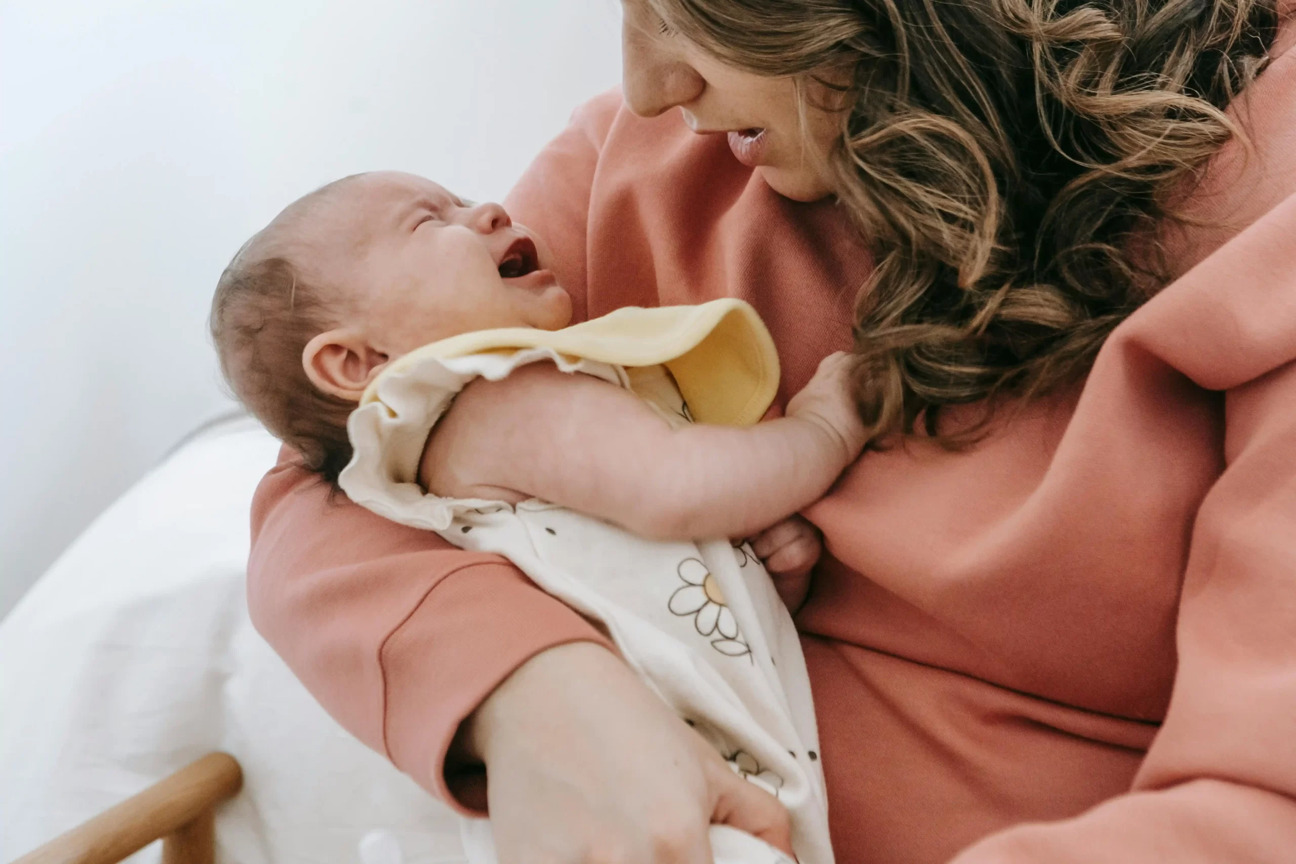 A mother wearing a pink sweater gently cradles and comforts her crying newborn baby in her arms.