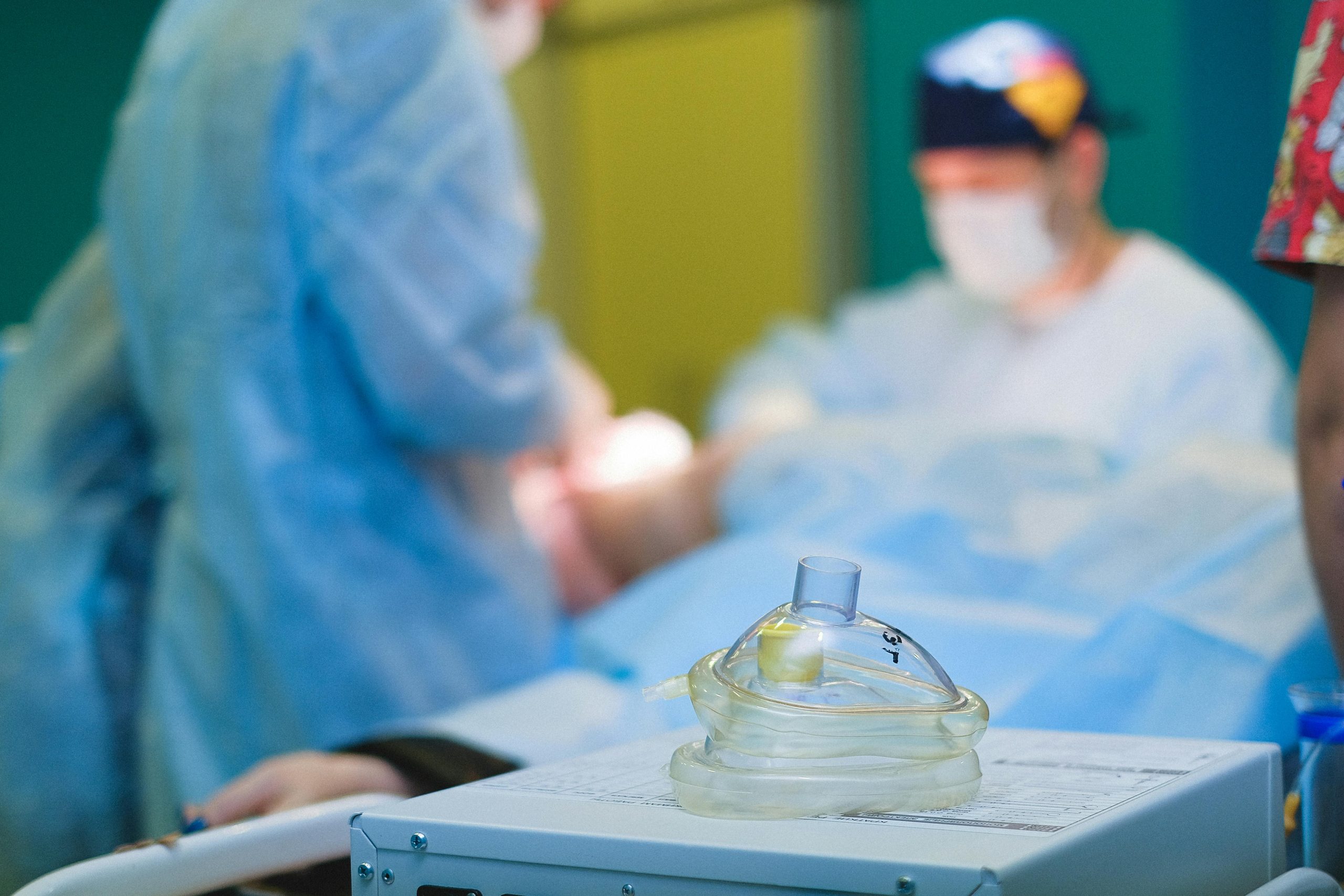 A clear anesthesia mask resting on a medical device in the foreground, with a blurred surgical team performing an operation in the background.