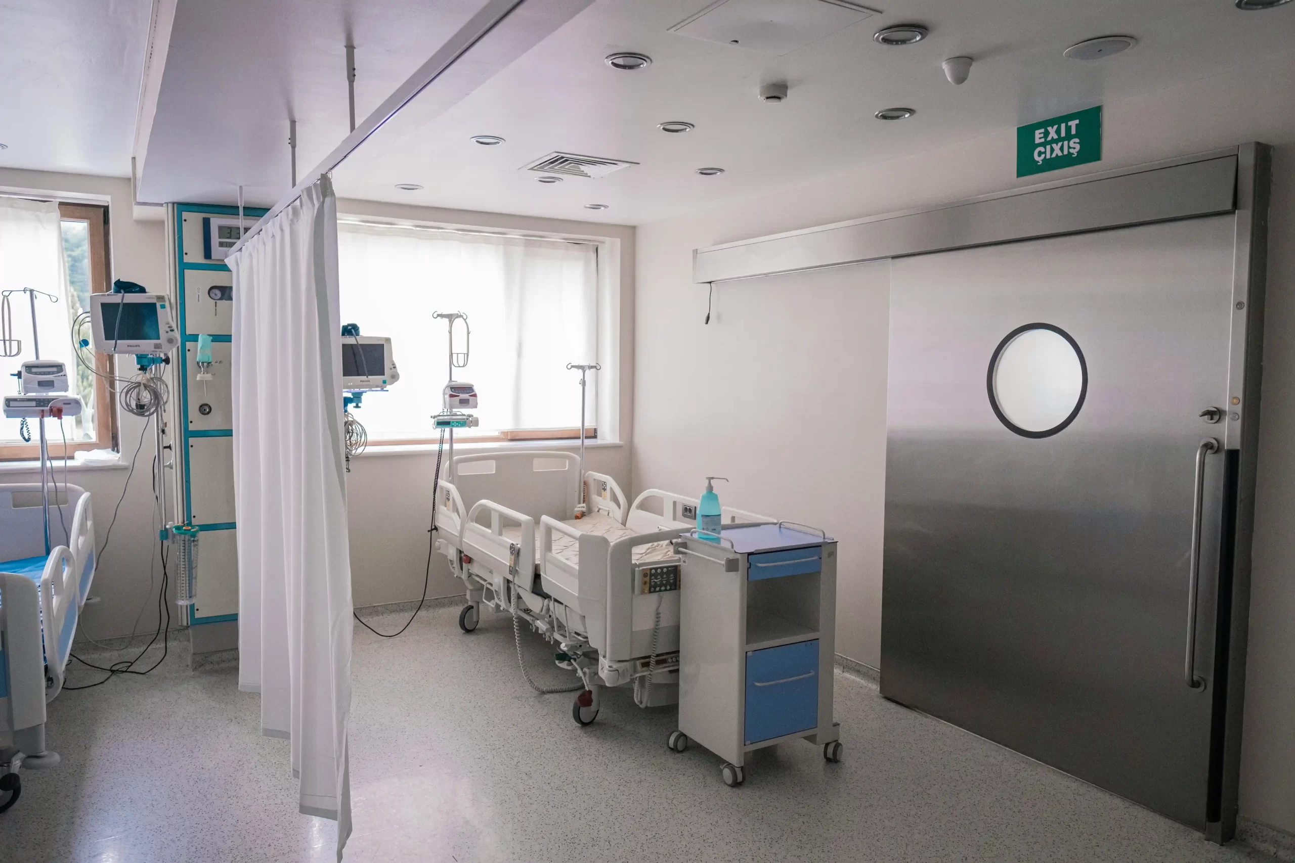 A clean, modern hospital room equipped with adjustable patient beds, medical monitors, a privacy curtain, and a sliding metal door.
