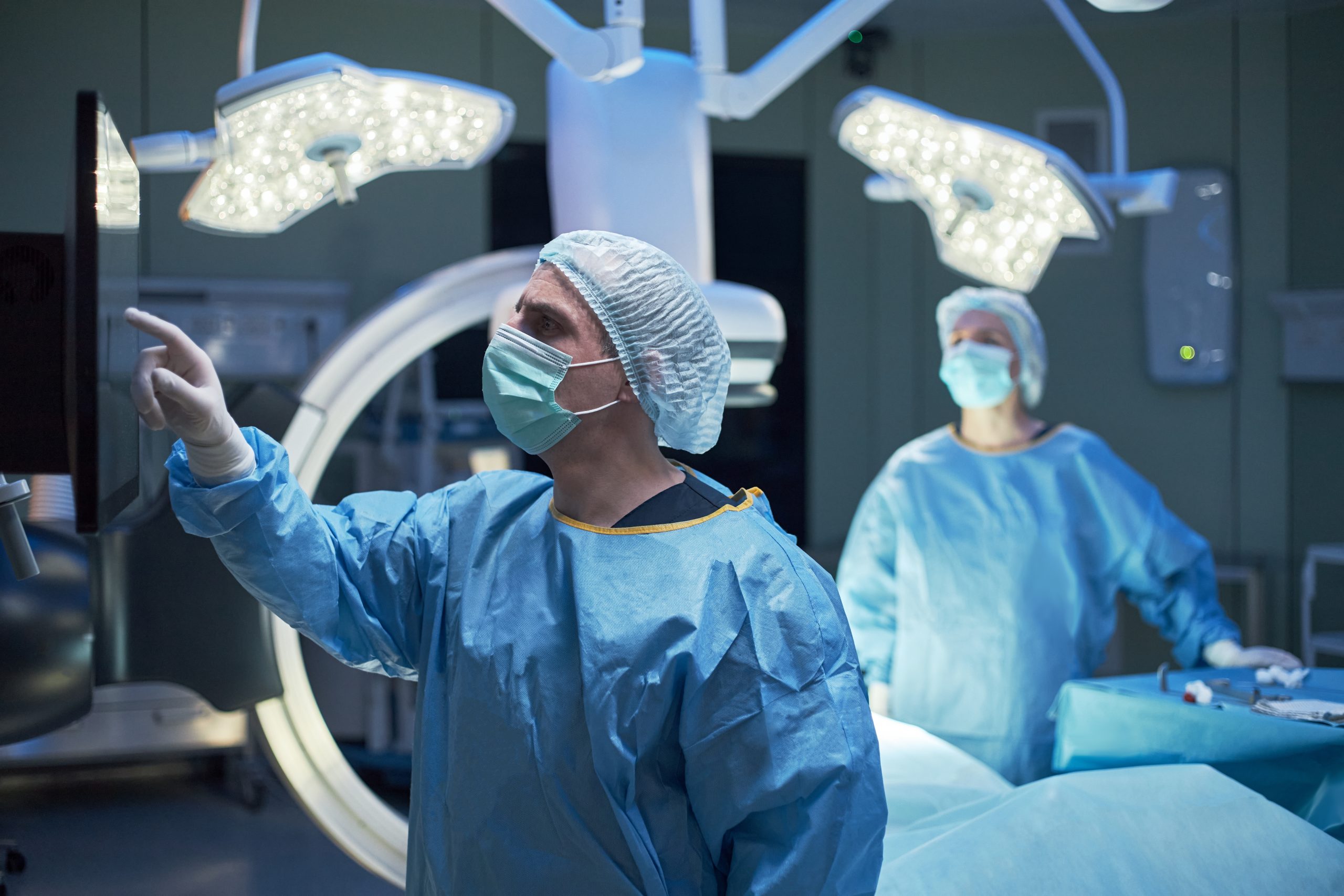 A male surgeon in blue scrubs and a face mask pointing at a touchscreen monitor in a modern operating room.