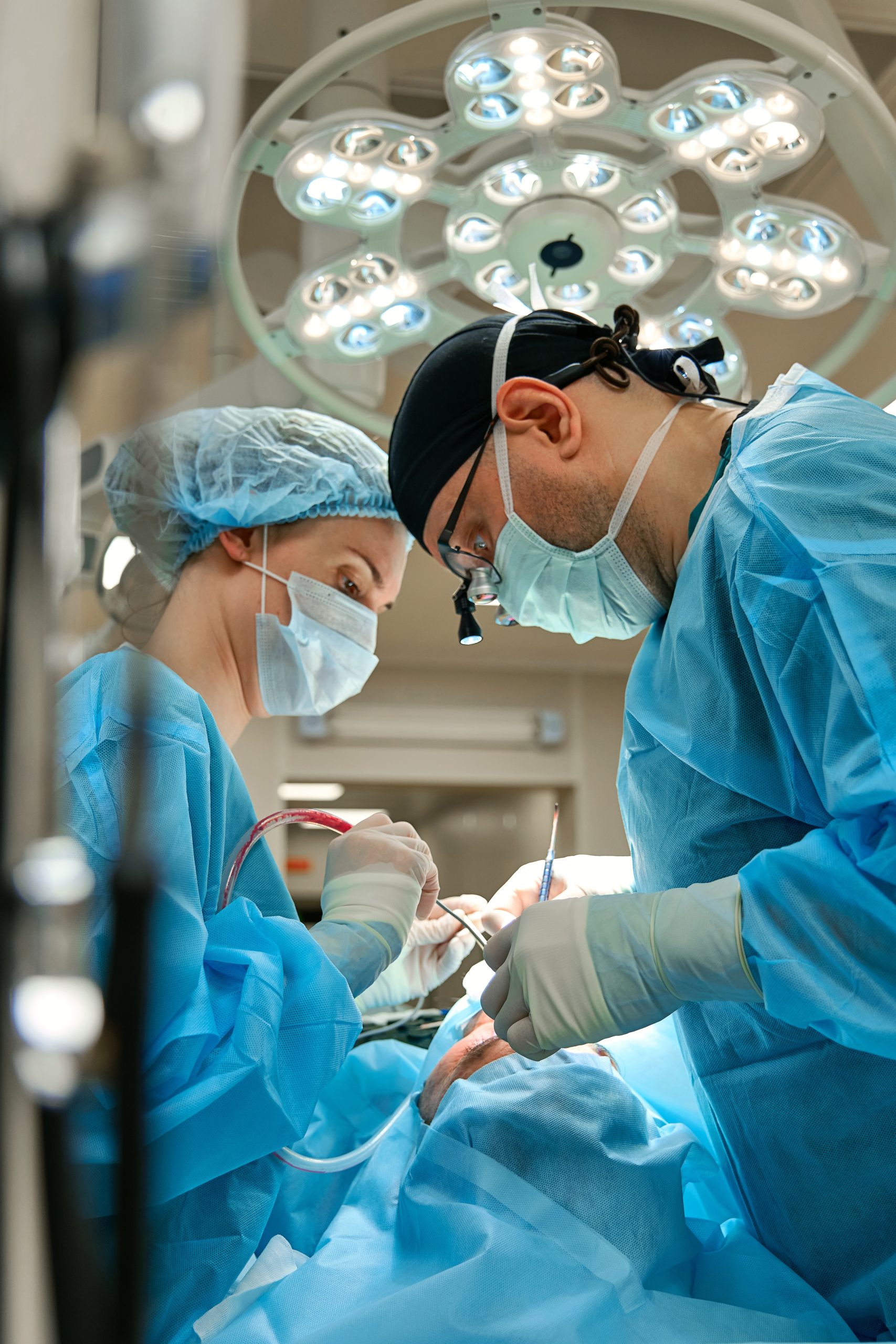 A surgeon with surgical loupes and an assistant performing a procedure under a large surgical light.